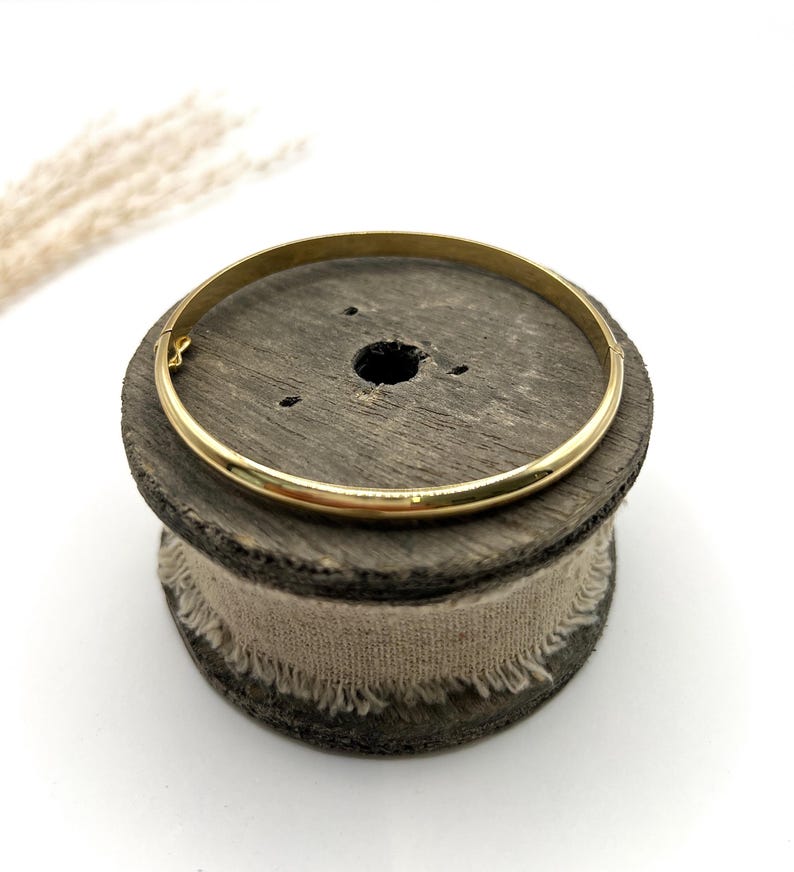 May include: A gold bangle bracelet with a simple design. The circular bracelet is made of metal and sits on a wooden spool with a beige fabric wrap. The background is white. The bracelet's diameter is approximately 6.5 cm.