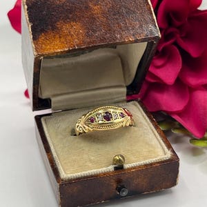 Peut inclure: Une bague en or avec une rangée centrale de trois petites pierres rouges alternant avec deux pierres claires, sertie dans un anneau en or texturé et allongé aux bords en filigrane, présentée dans un écrin à bijoux marron à charnière.
