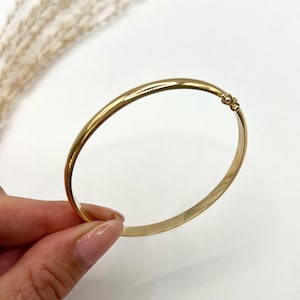 May include: A gold-colored bangle bracelet with a simple, elegant design. The bracelet has a smooth, polished surface and a small decorative clasp. The background is white with a hint of dried grass.