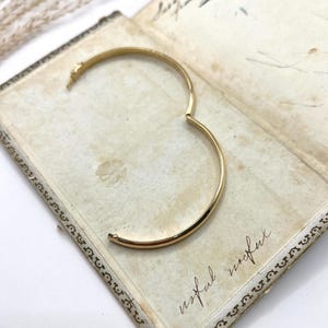 May include: A gold-coloured hinged bangle bracelet, open and resting on an antique book. The bracelet has a simple, rounded design. The book's pages are aged with handwritten script and the word "useful" is visible.