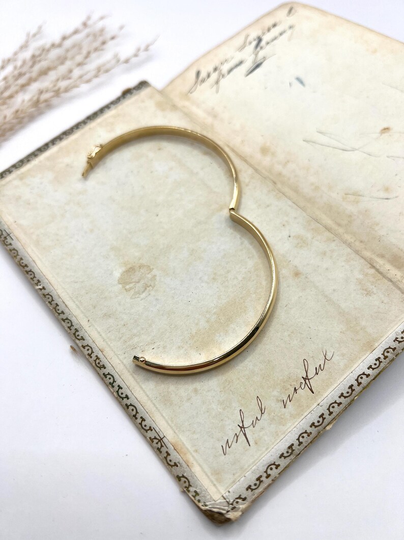 May include: A gold-coloured hinged bangle bracelet, open and resting on an antique book. The bracelet has a simple, rounded design. The book's pages are aged with handwritten script and the word "useful" is visible.