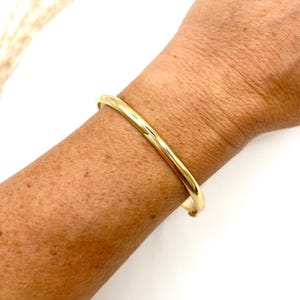 May include: A gold bangle bracelet. The bracelet is a simple, smooth band with a rounded profile. The image shows the bracelet on a person's arm, with the skin tone visible. The background is white.