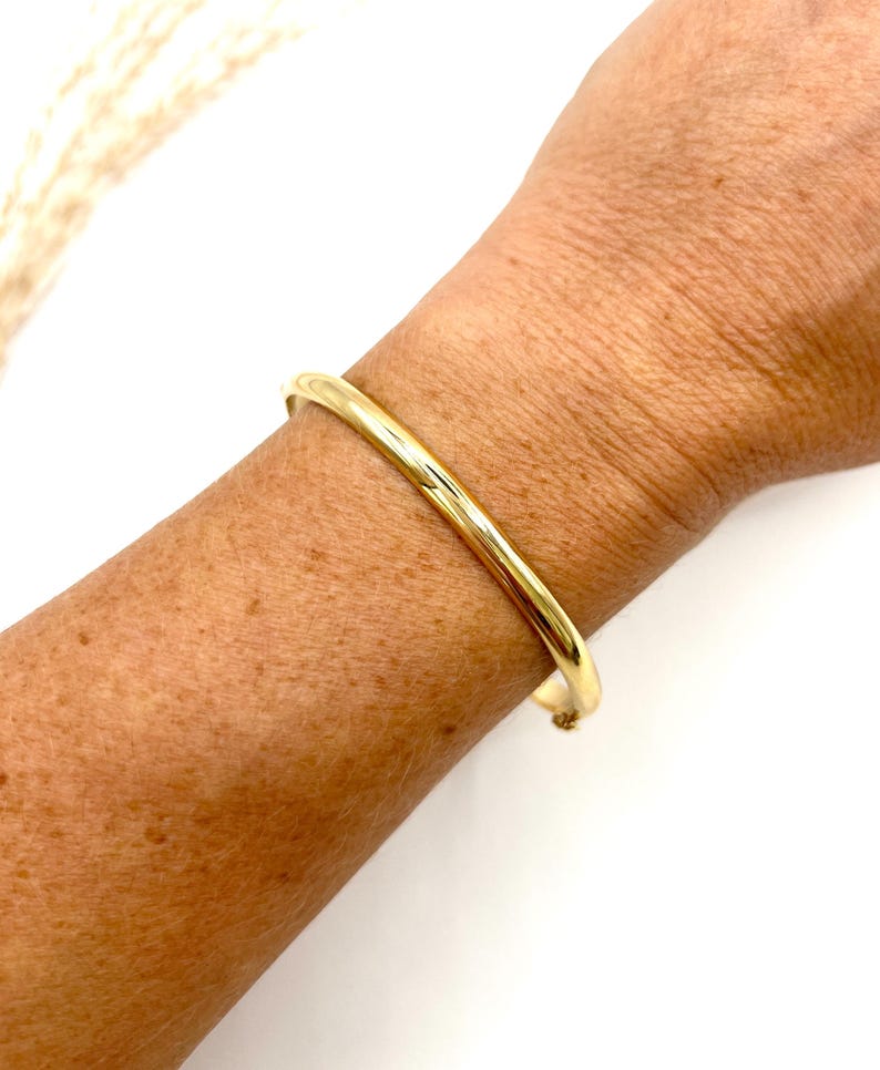 May include: A gold bangle bracelet. The bracelet is a simple, smooth band with a rounded profile. The image shows the bracelet on a person's arm, with the skin tone visible. The background is white.