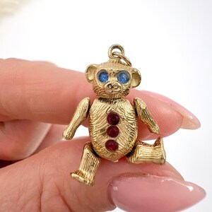 Op de afbeelding: Een gouden teddybeerbedel met blauwe ogen en rode edelsteenaccenten. De bedel ligt op een hand.