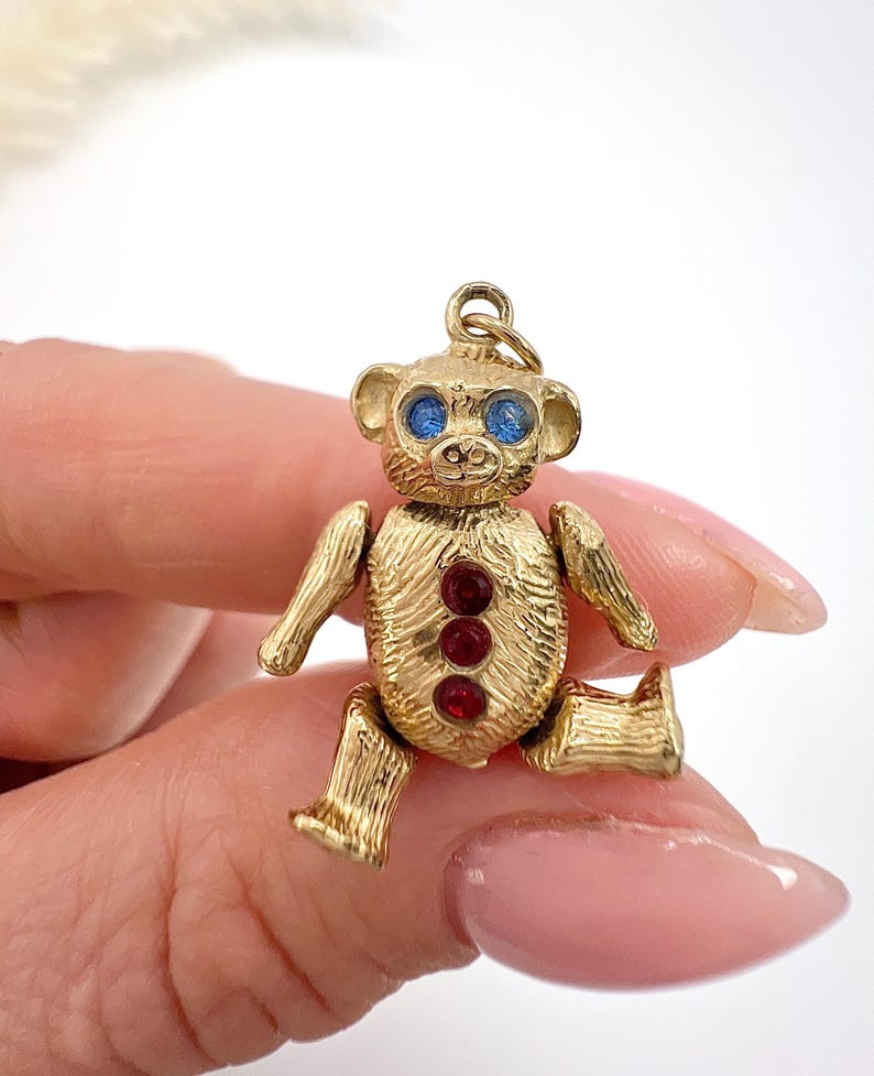 Op de afbeelding: Een gouden teddybeerbedel met blauwe ogen en rode edelsteenaccenten. De bedel ligt op een hand.