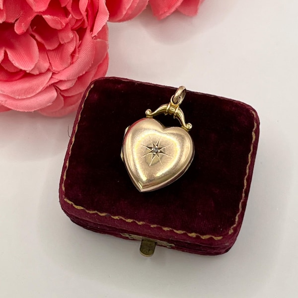 Könnte beinhalten: Ein herzförmiger Medaillon-Anhänger aus Gold mit einer kleinen, mittigen Stern-Gravur und einem winzigen eingesetzten Stein. Das Medaillon hat eine dekorative goldene Öse an der Oberseite und liegt auf einer kleinen, rechteckigen Schmuckschatulle aus bordeauxrotem Samt.