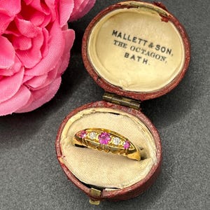 Op de afbeelding: Een gouden ring met roze en heldere edelstenen, gepresenteerd in een open, antieke, rode sieradendoos. De doos heeft een crèmekleurig interieur en de tekst "MALLETT & SON, THE OCTAGON, BATH." Op de achtergrond is een roze bloem te zien.