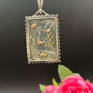Könnte beinhalten: Eine silberne Anhänger Halskette mit einem rechteckigen Anhänger mit einer detaillierten Naturszene. Der Anhänger hat einen silbernen Rahmen mit einem detaillierten Design, und die Szene umfasst Vögel, Blumen und Blätter in Gold- und Silbertönen.