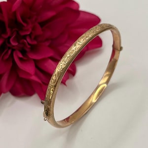 Può includere: Un bracciale rigido in metallo color oro caratterizzato da un intricato motivo floreale inciso sulla superficie esterna. Il bracciale circolare include un meccanismo di chiusura a cerniera su un lato, che permette di aprirlo e chiuderlo.