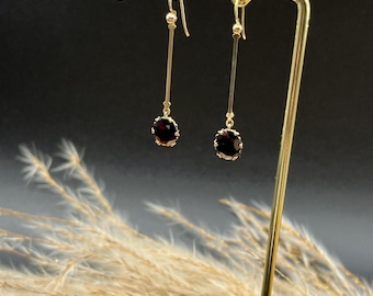 Könnte beinhalten: Goldfarbene Hängeohrringe mit dunkelroten Edelsteinen. Die Ohrringe hängen an einem goldenen Stab und werden auf einem goldenen Schmuckständer präsentiert. Der Hintergrund ist schwarz, mit getrocknetem Ziergras im Vordergrund.