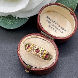 Peut inclure: Une bague en or avec une pierre rouge centrale entourée de petites pierres claires dans un sertissage géométrique, présentée dans un écrin rond en cuir rouge foncé avec le texte MALLETT & SON, THE OCTAGON, BATH sur la doublure crème.