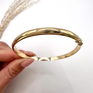May include: A gold-coloured bangle bracelet with a clasp. The bracelet has a smooth, polished surface and a rounded shape. The jewellery is displayed on a white background.