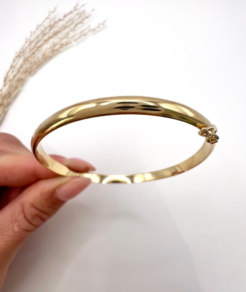 May include: A gold-coloured bangle bracelet with a clasp. The bracelet has a smooth, polished surface and a rounded shape. The jewellery is displayed on a white background.