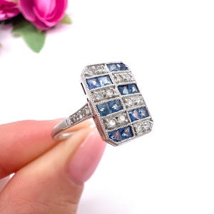 Könnte beinhalten: Ein rechteckiger Ring in Silberoptik mit einem geometrischen Gittermuster aus abwechselnd quadratisch geschliffenen blauen Saphiren und klaren weißen Diamanten. Die Ringschiene ist schmal und mit kleinen Diamantakzenten verziert.