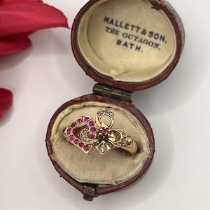 May include: A gold ring featuring a heart-shaped design set with small pink and clear stones, resting inside a round, hinged, dark red leather jewelry box. The interior lid of the box is lined with cream fabric and printed with MALLETT & SON, THE OCTAGON, BATH.