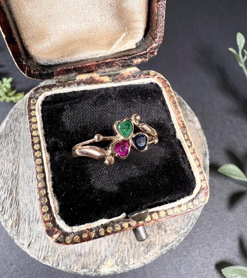 May include: A gold ring with three heart-shaped gemstones. The gemstones are green, pink, and blue.