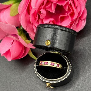 May include: A gold ring with alternating red and clear stones, displayed in an open black jewelry box. Pink roses are in the background, with a dark gray surface.