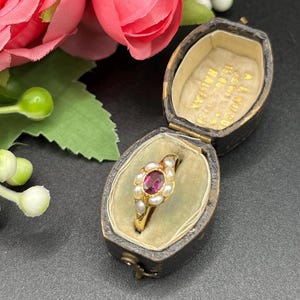 Może przedstawiać: Złoty pierścionek z owalnym, ciemnoróżowym kamieniem szlachetnym pośrodku, otoczonym małymi, białymi perłami. Pierścionek jest prezentowany w zabytkowym, czarnym skórzanym pudełku na biżuterię ze złotymi literami. Różowe kwiaty i zielone liście w tle.