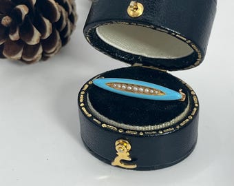 May include: A vintage brooch in a black jewelry box. The brooch features a light blue enamel center with a row of small pearls and gold-tone accents. The box is open, revealing a black velvet interior.