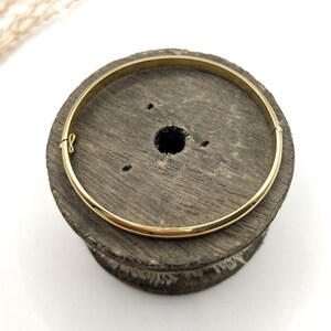 May include: A gold-coloured bangle bracelet with a clasp, resting on a dark wooden spool. The bracelet is a simple, circular design. The spool has a central hole and a rough, textured surface. The background is white.