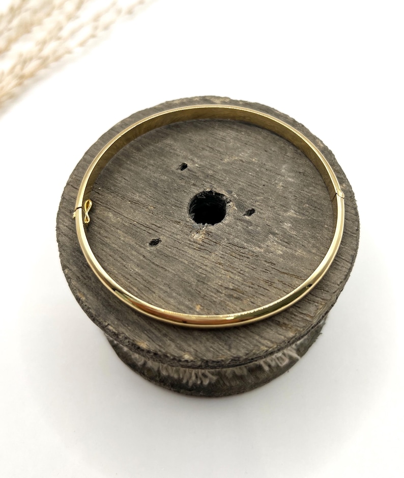 May include: A gold-coloured bangle bracelet with a clasp, resting on a dark wooden spool. The bracelet is a simple, circular design. The spool has a central hole and a rough, textured surface. The background is white.