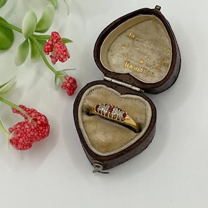 May include: A gold ring set with clear and red gemstones, presented in an open, heart-shaped brown leather jewellery box. The box has a beige interior. Red berries and green leaves are in the background.