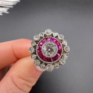 May include: A vintage-style ring featuring a central, round, clear diamond surrounded by a halo of small, vibrant red gemstones. The outer ring is composed of smaller, clear diamonds set in a silver-toned metal, creating a floral-like design.