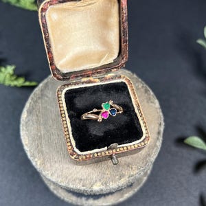 May include: A gold ring with three heart-shaped gemstones. The gemstones are green, pink, and blue. The ring is in a small, wooden box with a black velvet lining.