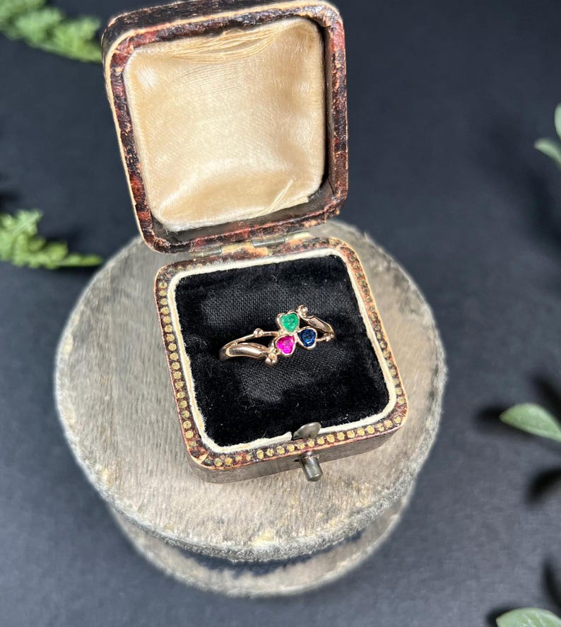 May include: A gold ring with three heart-shaped gemstones. The gemstones are green, pink, and blue. The ring is in a small, wooden box with a black velvet lining.