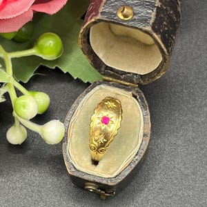 Może przedstawiać: Złoty pierścionek z centralnym czerwonym kamieniem szlachetnym, osadzonym w gwiazdowym wzorze. Pierścionek jest prezentowany w antycznym, ciemnym skórzanym pudełku na biżuterię z aksamitnym wnętrzem. Opaska pierścionka ma skomplikowane detale.