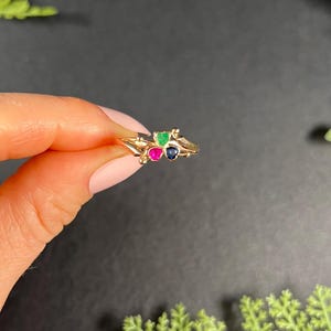 May include: A gold ring with three small gemstones: a green emerald, a pink ruby, and a blue sapphire.
