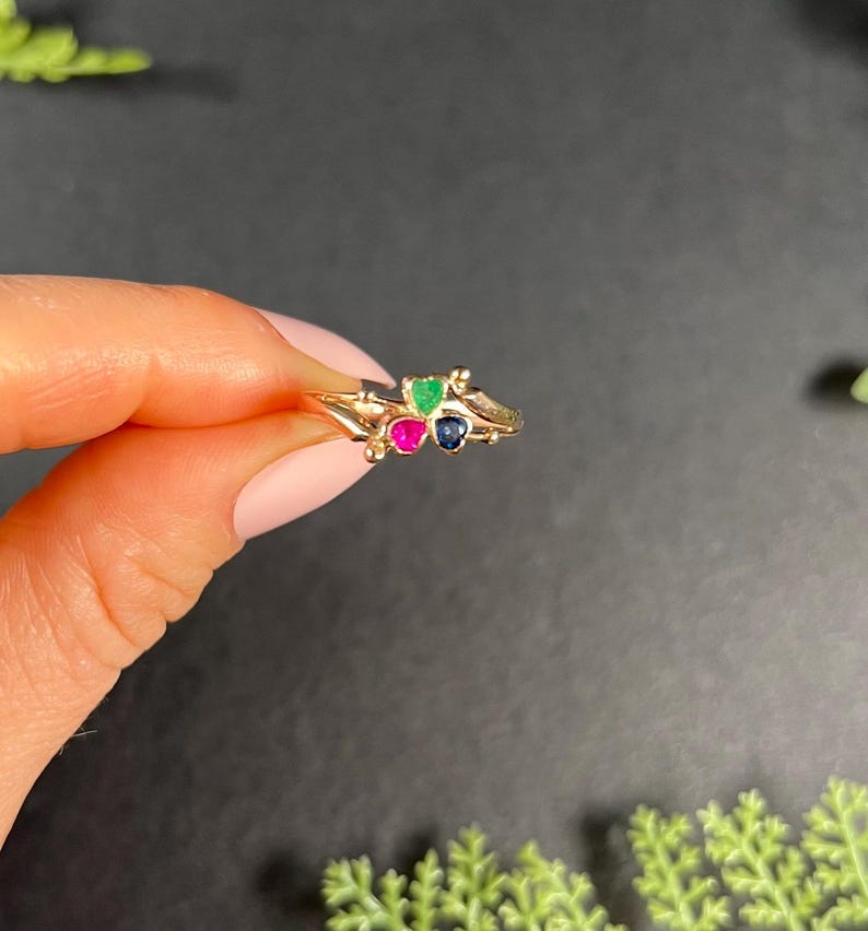 May include: A gold ring with three small gemstones: a green emerald, a pink ruby, and a blue sapphire.