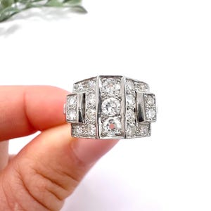 May include: A silver-toned ring featuring a rectangular face set with multiple round, clear, faceted stones. The central vertical row contains three prominent stones, flanked by smaller stones arranged in a geometric, stepped pattern.