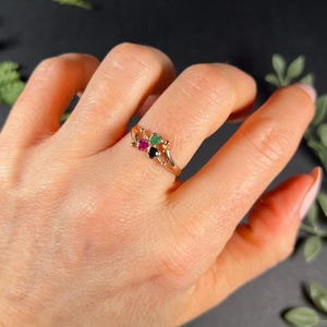 May include: A gold ring with three heart-shaped gemstones. The gemstones are green, red, and black.