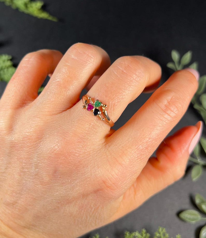 May include: A gold ring with three heart-shaped gemstones. The gemstones are green, red, and black.