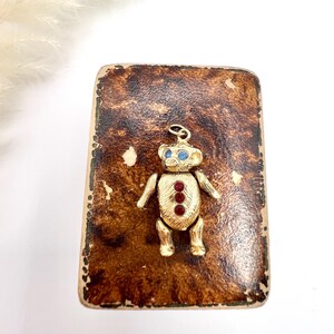 Op de afbeelding: Een gouden teddybeerbedel met blauwe ogen en rode edelsteenaccenten, gepresenteerd op een bruin, getextureerd oppervlak.