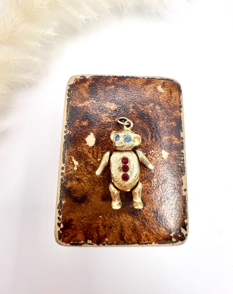 Op de afbeelding: Een gouden teddybeerbedel met blauwe ogen en rode edelsteenaccenten, gepresenteerd op een bruin, getextureerd oppervlak.