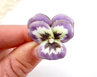 以下が含まれることがあります： パンジーの形をした花のブローチのクローズアップ。ブローチは、白と緑のアクセントが付いた紫色の花びらと、中央に小さな輝く宝石が特徴です。ブローチは手で持たれています。