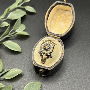 Könnte beinhalten: Ein goldener Vintage-Ring mit einem zentralen dunklen Stein, umgeben von kleinen Perlen, präsentiert in einer offenen, antiken schwarzen Schmuckschatulle mit cremefarbenem Innenraum. Der Ring hat florale Akzente. Die Schatulle hat Text.
