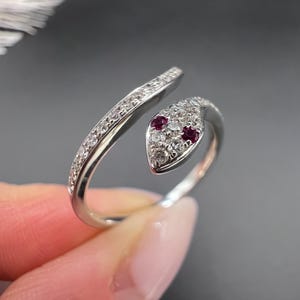 May include: A silver snake ring with a curved design. The ring's head features two red gemstones as eyes and is surrounded by small, clear gemstones. The body of the ring is also adorned with clear gemstones.