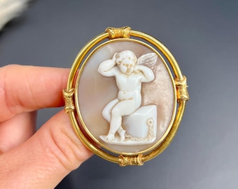 May include: A gold-toned oval cameo brooch featuring a carved shell cameo of a winged cherub sitting on a block. The cherub is carved in white against a pink shell background.