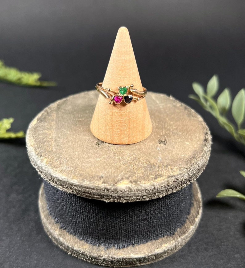 May include: A gold ring with three heart-shaped gemstones. The gemstones are green, pink, and black.