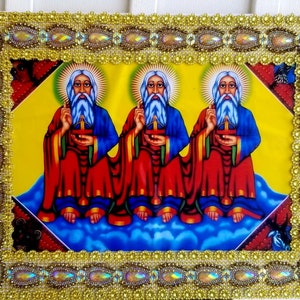 May include: A framed religious icon depicting three figures with long white beards and halos. They are wearing red and blue robes and holding a cross. The frame is decorated with gold and iridescent beads.