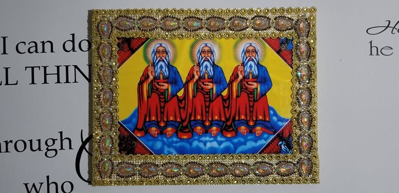 May include: A framed religious icon with three figures in red and blue robes, sitting on a cloud with a yellow background. The frame is gold with a decorative border.