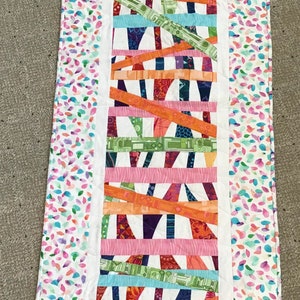 May include: A rectangular quilted table runner with a colorful abstract design. The center features horizontal strips of various patterned fabrics in orange, green, blue, and pink. The border has a white background with a pattern of colorful leaf shapes.