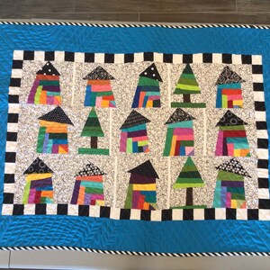 May include: A colorful quilt featuring houses and trees. The houses are made of stacked, multicolored rectangles, and the trees are green. The quilt has a black and white checkered border and a blue quilted edge.