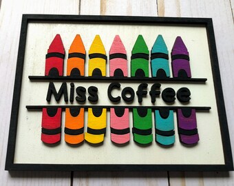 May include: A wooden sign with a black frame and a white background. The sign features a row of colorful crayons with the text "Miss Coffee" in black letters.