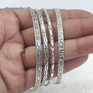 Puede incluir: Conjunto de cuatro pulseras de plata con diferentes diseños en relieve. Las pulseras están apiladas una encima de la otra y tienen un acabado brillante.