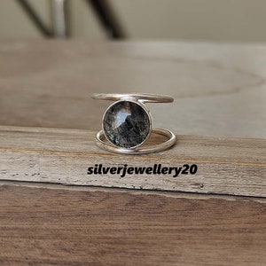 Könnte beinhalten: Ein silberner Ring mit einem schwarzen und klaren Edelstein, der in einem Doppelband-Design gefasst ist. Der Ring liegt auf einer Holzoberfläche.