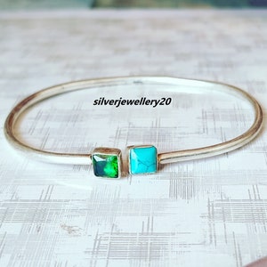 May include: A silver bangle bracelet with two square-shaped gemstones. The gemstone on the left is green and the gemstone on the right is turquoise.